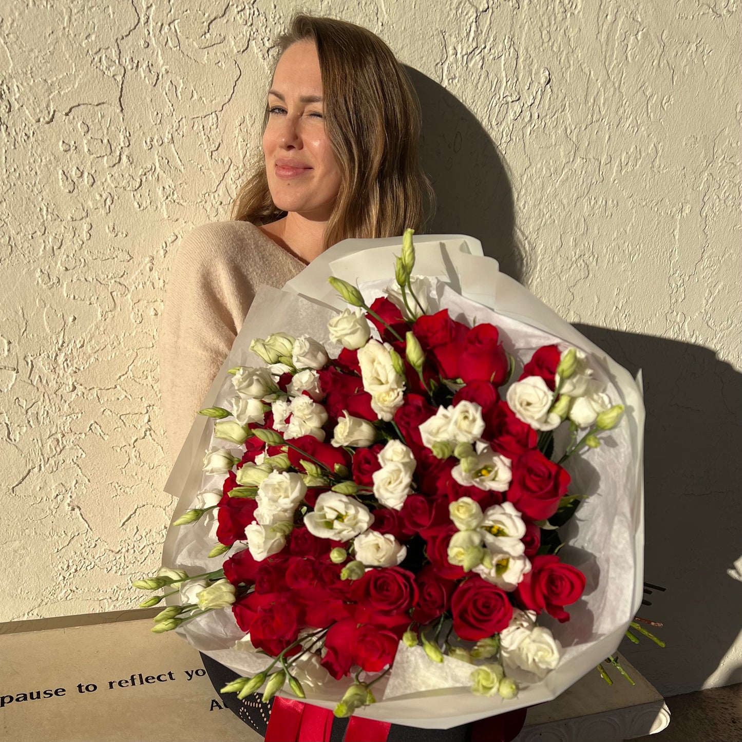 A gorgeous woman holding perfect bouquet of red roses and white lisianthuses sits on a bench and