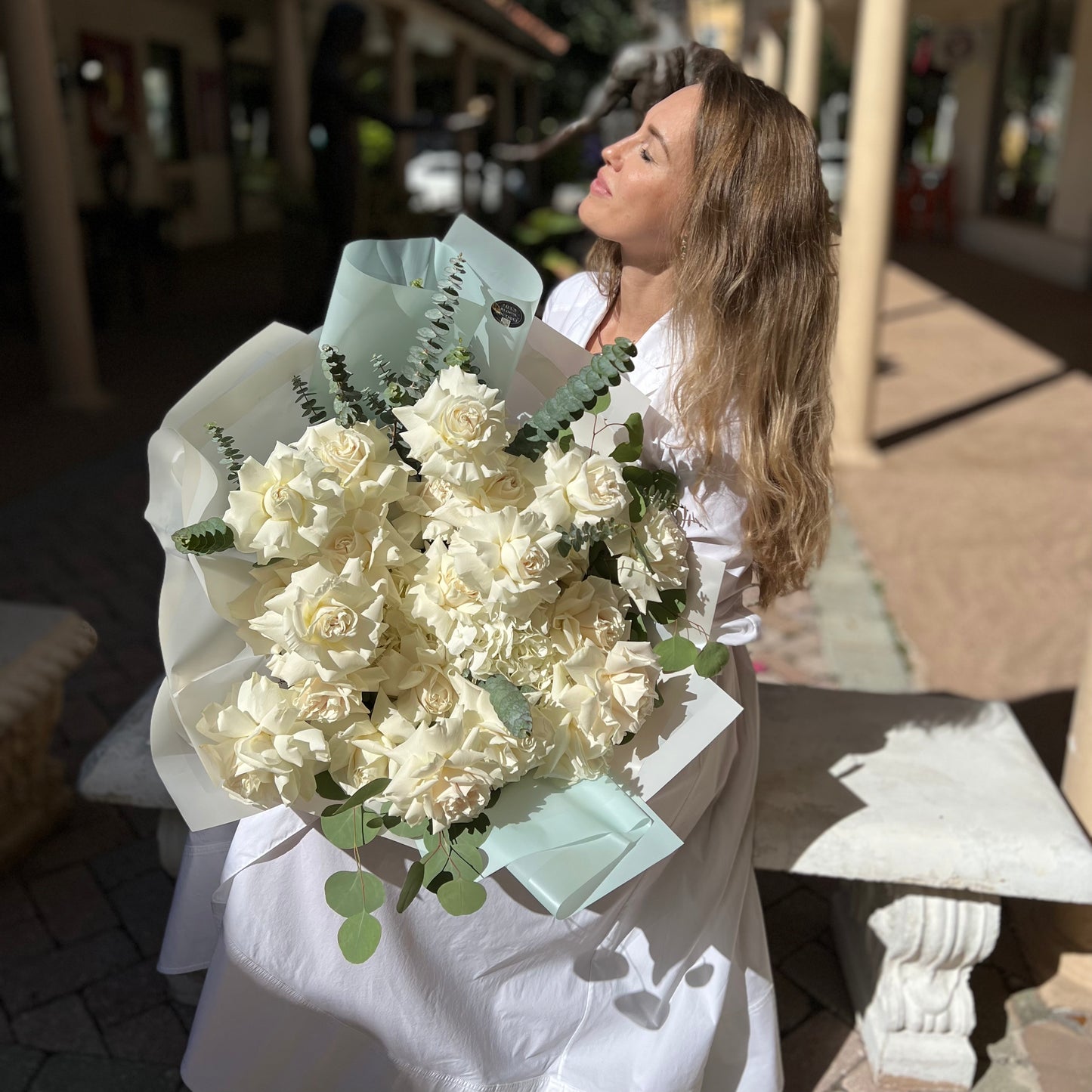 White roses, hydrangea and a dash of greenery in a bouquet held by a woman sitting on a concrete bench 